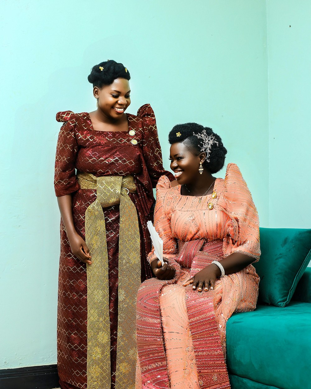 Two women in coordinated maroon and coral gomesi, laughing together