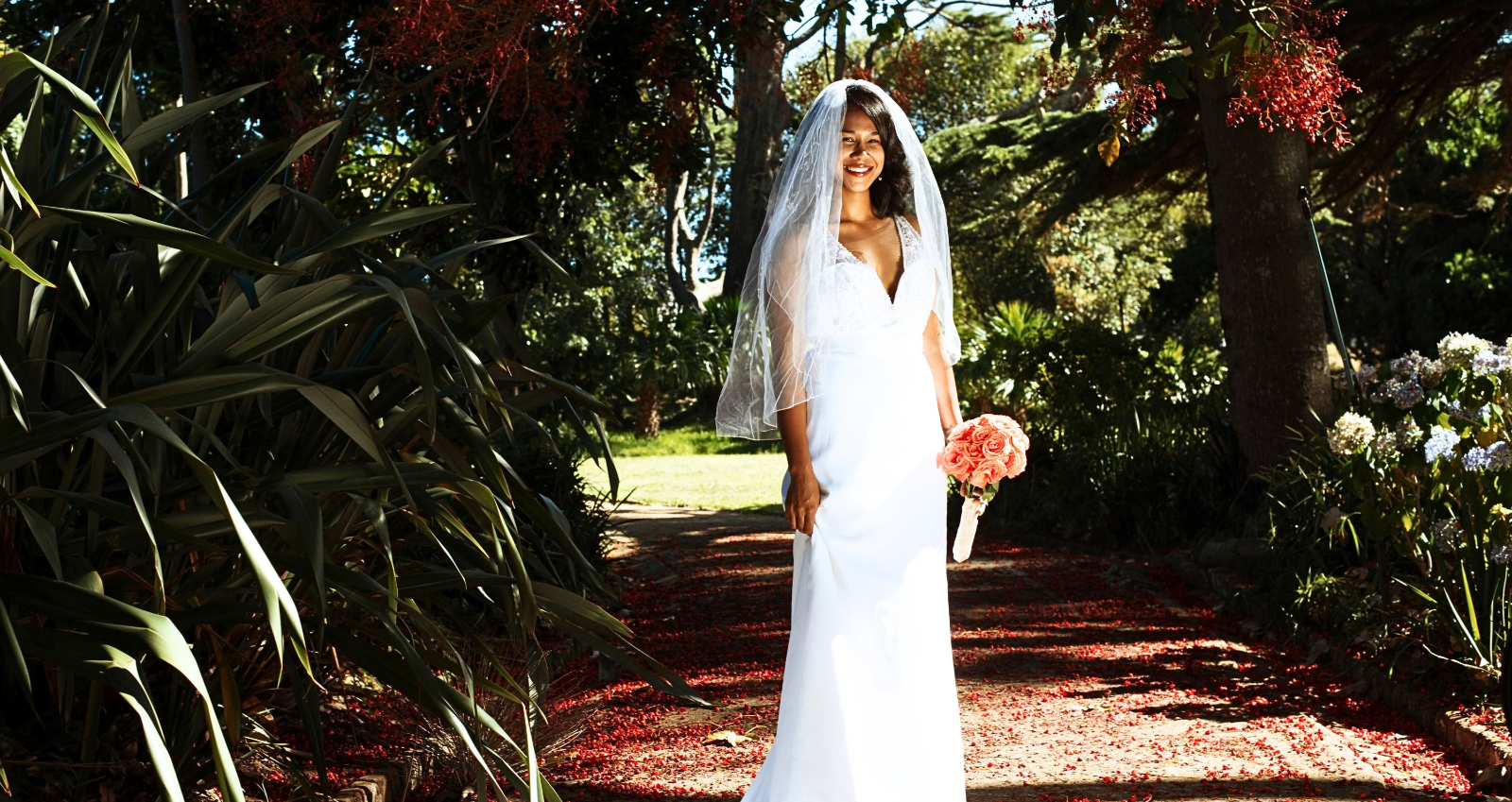 A smiling bride in a white gown and veil holding coral roses on a petal-strewn garden path
